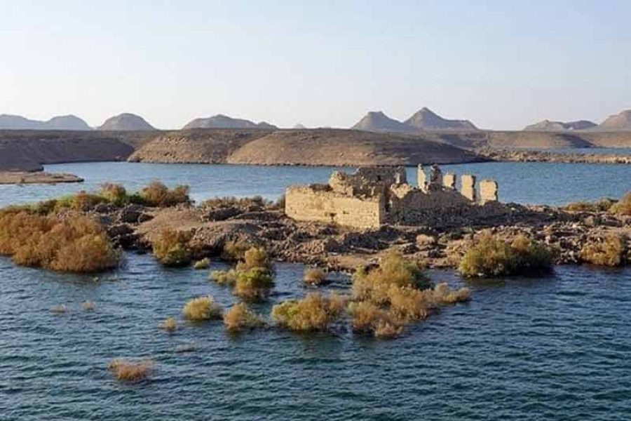 The partially submerged ancient fortress of Kasr Ibrim, an archaeological island, rising from the tranquil blue waters of Lake Nasser, with the vast lake stretching into the distance