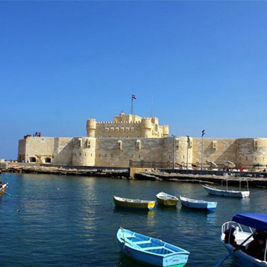 The imposing Qaitbay Fortress standing proudly on the coast of Alexandria, with numerous fishing boats and smaller vessels visible in the blue Mediterranean waters below