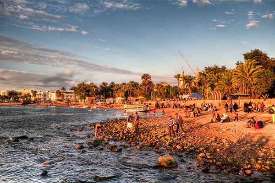 azure waters of Aqaba's Red Sea beach, with visitors relaxing on the sand and the distinctive, rugged mountains of Jordan