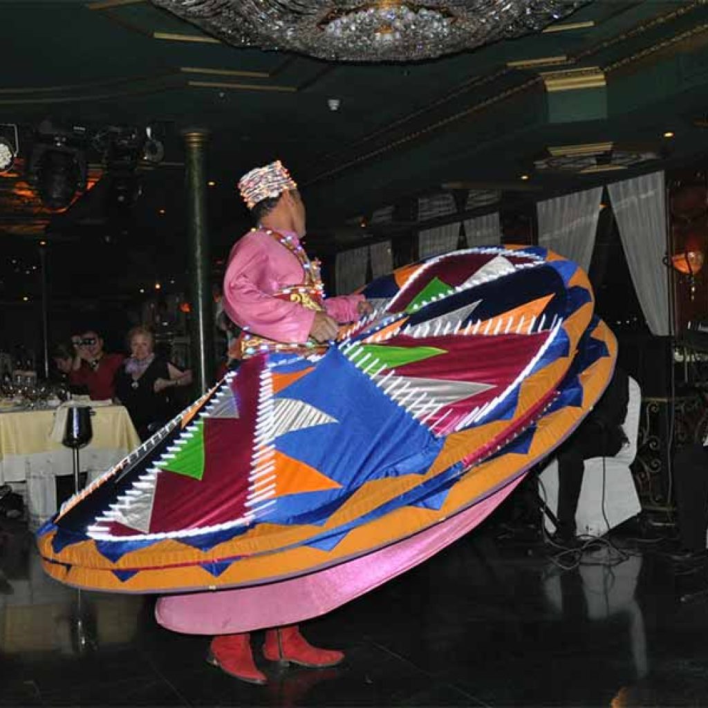 A male Tanoura dancer in a vibrant, multi-layered skirt spinning gracefully on a dinner cruise boat