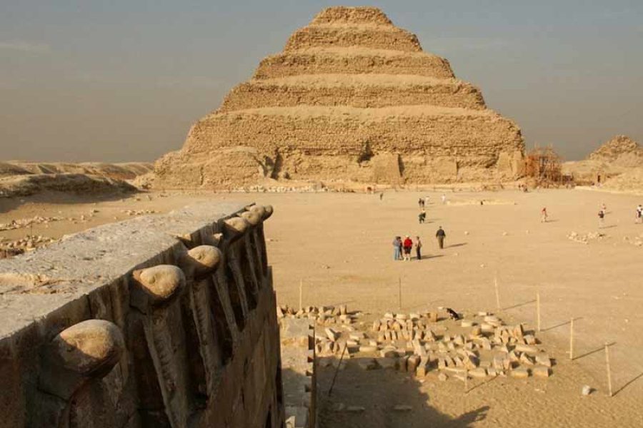 The ancient, multi-tiered silhouette of the Djoser Step Pyramid at Saqqara, showing its distinct steps rising under a clear desert sky, symbolizing the dawn of monumental stone architecture