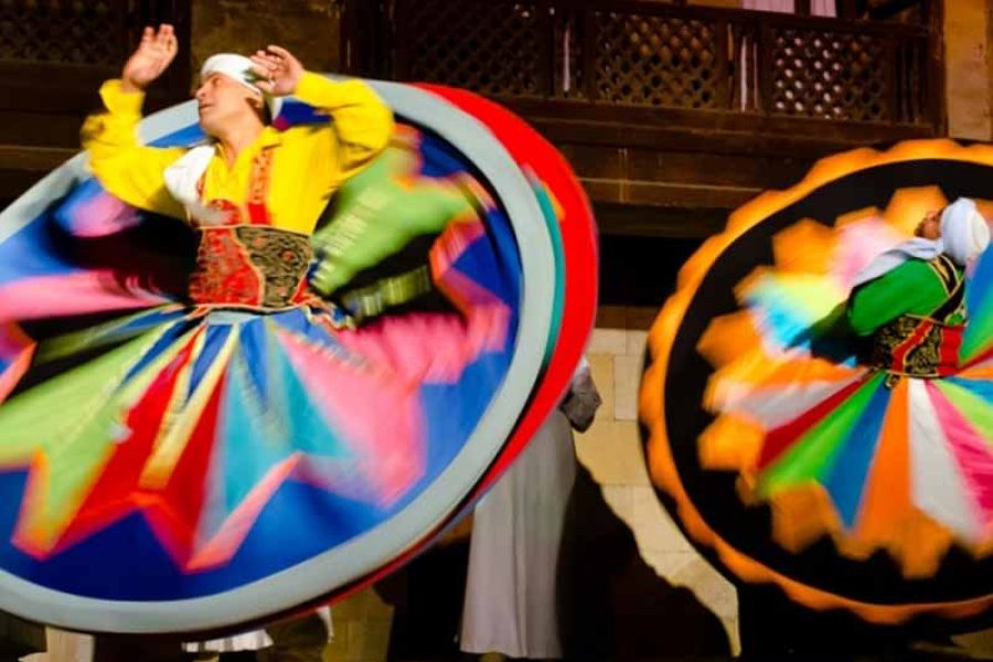 A male Tanoura dancer in a vibrant, multi-layered skirt spinning gracefully on stage within the historic, illuminated courtyard of Wekalet El Ghouri in Old Cairo