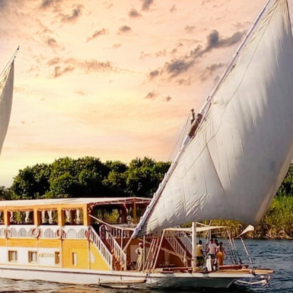 An elegant, traditional Dahabiya sailboat with large, white canvas sails gently gliding on the Nile River