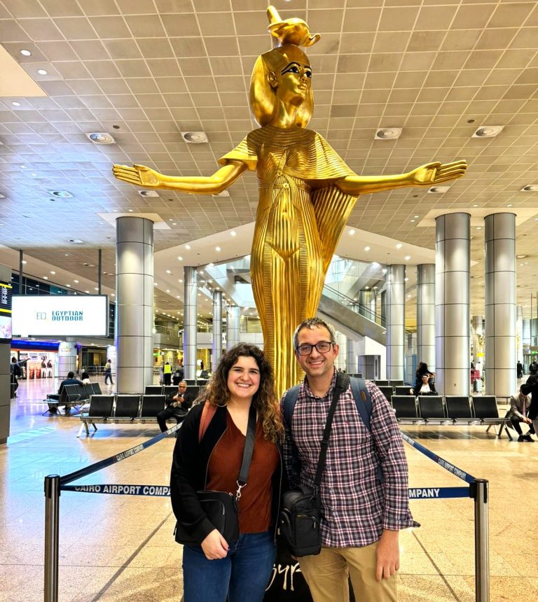 A happy couple smiling and holding hands in the busy terminal of Cairo International Airport, looking excited for their upcoming trip to Egypt