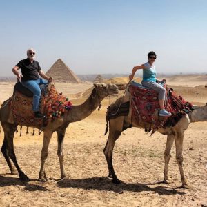 La photo montre deux personnes à dos de chameau dans le quartier des pyramides, avec leur propriétaire à leurs côtés. Derrière elles se trouvent deux pyramides de Gizeh.