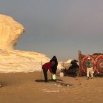 Campement dans le désert blanc égyptien avec des tentes, des personnes et un véhicule devant une formation rocheuse blanche.