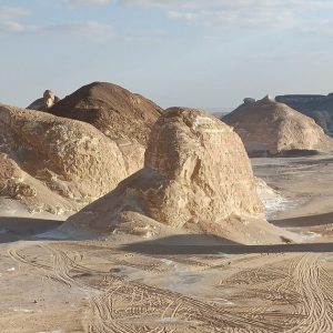 Formations rocheuses uniques blanches et brunes avec des traces de pneus dans le sable sous un ciel lumineux, suggérant un paysage désertique.
