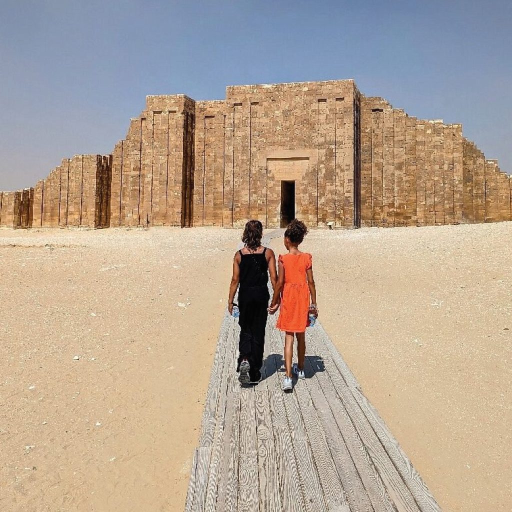 Deux enfants marchant main dans la main sur une passerelle en bois, en direction d'une structure monumentale en pierre dans le désert sous un ciel dégagé.