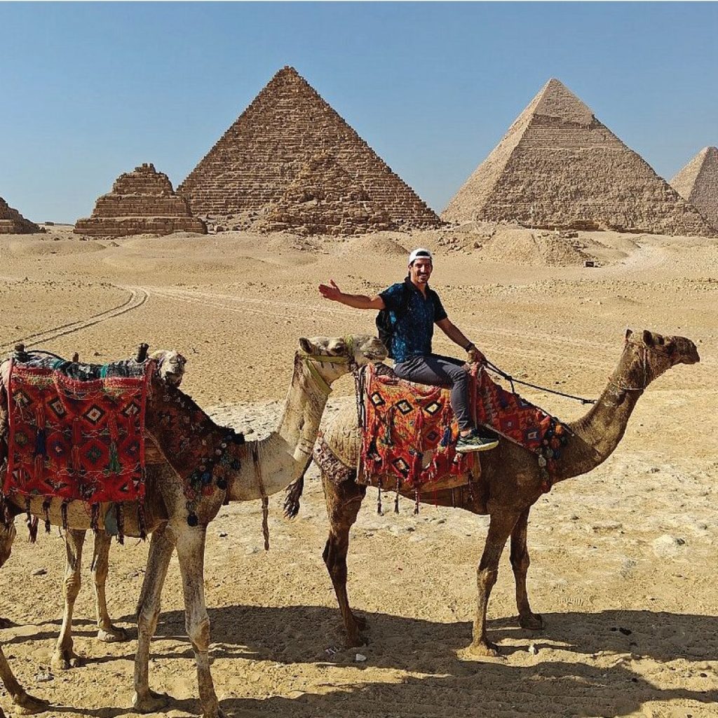 Un homme souriant monte un chameau décoré de tapis traditionnels, avec les pyramides de Gizeh en arrière-plan dans le désert égyptien sous un ciel clair.