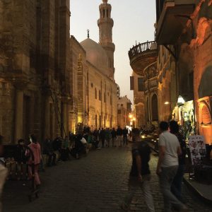 Rue pavée animée du Caire islamique au crépuscule, avec des passants, des bâtiments historiques éclairés, une mosquée à dôme et minaret en arrière-plan.