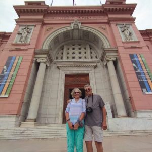 Un couple âgé, une femme et un homme, posent devant la façade rose-rouge et à colonnes du Musée égyptien. Le bâtiment possède une grande entrée arquée avec des sculptures ornées au-dessus. 