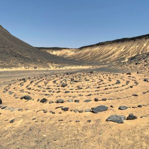 Une formation artistique de pierres sombres disposées en spirale sur le sable doré du désert. La spirale occupe le premier plan et le milieu de l'image, avec des cercles concentriques de roches menant à un centre. En arrière-plan, le paysage désertique est dominé par des collines et des montagnes arides, certaines de couleur foncée et d'autres plus claires, presque blanches, sous un ciel bleu clair et dégagé. Le terrain est plat et sablonneux autour de la spirale, parsemé de quelques petites roches.