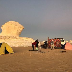 Un campement de nuit dans un paysage désertique avec des formations rocheuses crayeuses blanches emblématiques. Au premier plan et au centre, plusieurs personnes sont rassemblées autour d'un SUV blanc garé, sur lequel est accroché un tapis coloré à motifs géométriques. Deux tentes igloo, l'une jaune et bleue sur la gauche, et l'autre rose à droite, sont montées sur le sable. Le sol est couvert de sable clair et de traces de pas. À l'arrière-plan, des formations rocheuses blanches distinctives se dressent, la plus grande étant une grande masse sculpturale sur la gauche. Le ciel au-dessus est d'un bleu foncé, suggérant le crépuscule ou l'aube.