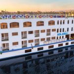 Un grand bateau de croisière sur le Nil blanc et bleu, nommé "SOLARIS I" sur sa proue, naviguant sur une étendue d'eau calme. Le bateau a plusieurs ponts avec de nombreuses fenêtres, des balcons et une rangée de chaises longues bleues sur le pont supérieur.