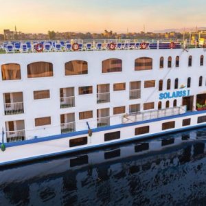 Un grand bateau de croisière blanc et bleu, nommé "SOLARIS I" sur sa proue, naviguant sur une étendue d'eau calme. Le bateau a plusieurs ponts avec de nombreuses fenêtres, des balcons et une rangée de chaises longues bleues sur le pont supérieur.