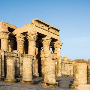 Une vue des ruines majestueuses du Temple de Kom Ombo en Égypte, baignées par une lumière claire. Les imposantes colonnes de pierre calcaire, certaines encore debout avec leurs chapiteaux ornés et des hiéroglyphes sculptés, dominent le cadre. Le temple, de couleur sable, présente des murs et des piliers partiellement effondrés mais conserve une grande partie de sa structure ancienne, témoignant de sa grandeur passée. Le sol autour est rocailleux et plat, et le ciel est d'un bleu clair et uniforme.