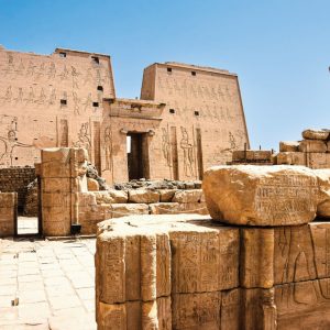 Une vue des ruines antiques du temple d'Edfou en Égypte sous un ciel bleu clair. Au centre de l'image, l'entrée principale du temple avec son imposant pylône décoré de hiéroglyphes et de grandes figures sculptées est visible. Au premier plan, des blocs de pierre tombés et des fûts de colonnes brisés, également couverts d'inscriptions, s'étendent sur un sol pavé de dalles. L'architecture est massive et de couleur sable, typique des temples égyptiens antiques, et une colonne solitaire se dresse à droite.