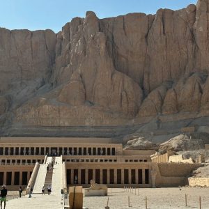 Une vue large du Temple mortuaire d'Hatchepsout en Égypte, s'étendant à la base d'imposantes falaises rocheuses ocres. Le temple est caractérisé par ses terrasses successives et ses longues rampes centrales qui montent vers l'édifice principal. Les façades du temple sont rythmées par des colonnades sombres. Au premier plan, un large chemin pavé mène vers le temple, avec quelques visiteurs visibles. Le sol autour du temple est fait de sable et de terre, et le ciel est d'un bleu clair éclatant.