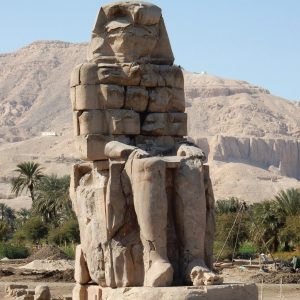 Une statue massive et ancienne des Colosses de Memnon se dresse sous un ciel clair, avec des montagnes arides en arrière-plan. La statue, fortement érodée et de couleur claire, représente une figure assise, probablement un pharaon, et est composée de plusieurs blocs de pierre empilés. Le sol autour de la statue est terreux et rocailleux. À gauche et à droite, on peut apercevoir de la verdure, notamment des palmiers, indiquant la présence d'une oasis ou d'une zone plus fertile à proximité des montagnes.