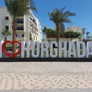 Une grande enseigne blanche tridimensionnelle sur une base noire qui épelle "I ❤️ HURGHADA" (J'❤️ HURGHADA), avec un cœur rouge vif remplaçant le mot "love". L'enseigne est placée sur une place pavée de dalles de couleurs claires, sous un ciel bleu clair. En arrière-plan, on voit des bâtiments modernes de couleur claire, typiques d'un complexe hôtelier ou résidentiel, ainsi que des palmiers.
