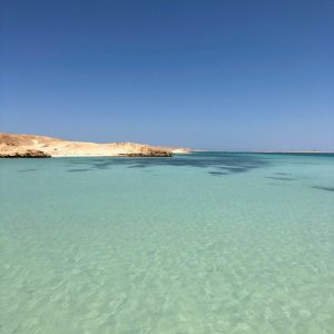 Une étendue d'eau cristalline aux teintes turquoise et bleu clair s'étendant sous un ciel azur dégagé. Le premier plan est dominé par les eaux peu profondes qui révèlent le fond sablonneux. À l'horizon, une côte désertique et sablonneuse avec des formations rocheuses de couleur ocre s'étend, se jetant dans la mer. L'eau devient plus foncée vers l'horizon, indiquant une profondeur croissante.