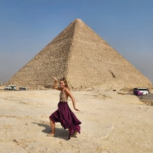 Une femme blonde vêtue d'une jupe pourpre virevoltante et d'un haut à motifs se tient sur un terrain rocailleux, les bras levés dans une pose de danse devant la Grande Pyramide de Gizeh. Le ciel est dégagé et on peut voir des bus et d'autres véhicules garés à l'arrière-plan.