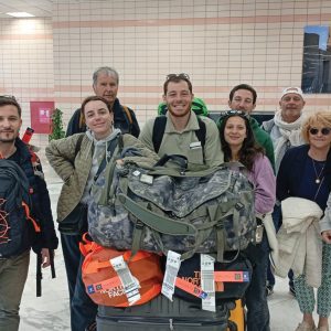 Huit personnes, cinq hommes et trois femmes, posent ensemble à l'intérieur d'un terminal d'aéroport. Ils ont plusieurs grands sacs de voyage et valises à leurs pieds. L'arrière-plan présente un mur carrelé beige et blanc, un panneau publicitaire en arabe et des indications aéroportuaires. Une plante en pot est visible sur la gauche.