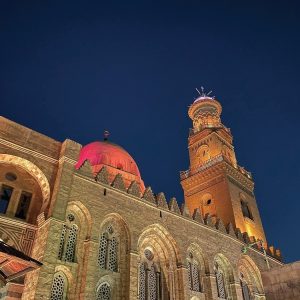 Une mosquée de couleur sable avec un grand dôme rose illuminé et un minaret éclairé est photographiée de bas en haut durant la nuit. Le ciel est bleu foncé avec quelques étoiles visibles. La mosquée a de nombreuses fenêtres à arc et des détails architecturaux complexes. Une partie d'un autre minaret est visible à l'extrême droite.
