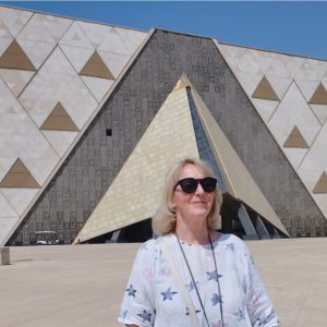 Une femme souriante en lunettes de soleil posant devant l'entrée du Grand Musée Égyptien au Caire, avec son architecture moderne en forme de pyramide.