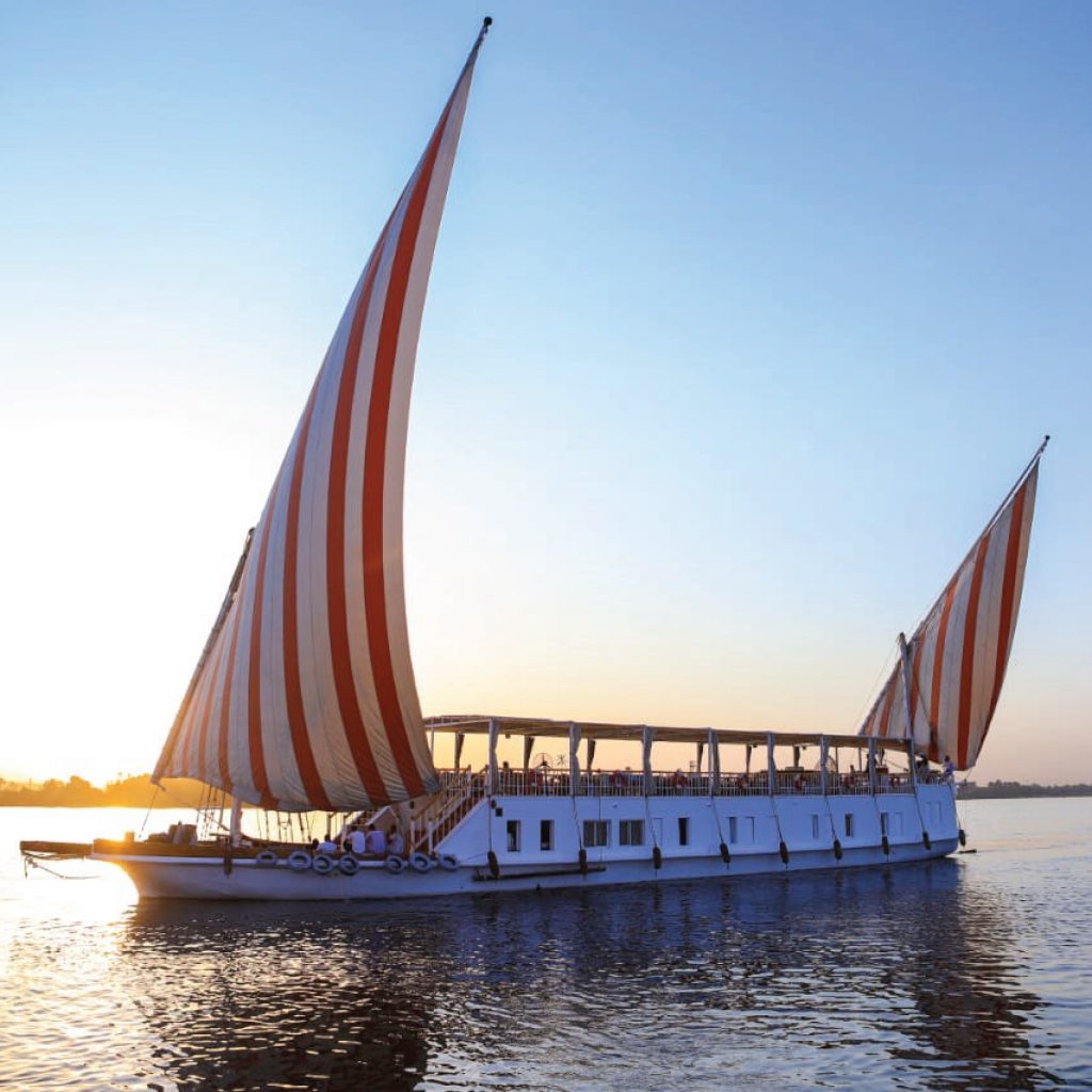 Une Croisière en Dahabyia , un grand voilier égyptien blanc avec deux voiles rayées de rouge et blanc, navigue sur le Nil au coucher du soleil. Le soleil, à gauche, se reflète sur l'eau et le ciel est un mélange de bleu clair et de teintes jaunes et roses. Les rives du fleuve sont visibles à l'arrière-plan.