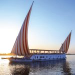 Une Croisière en Dahabyia , un grand voilier égyptien blanc avec deux voiles rayées de rouge et blanc, navigue sur le Nil au coucher du soleil. Le soleil, à gauche, se reflète sur l'eau et le ciel est un mélange de bleu clair et de teintes jaunes et roses. Les rives du fleuve sont visibles à l'arrière-plan.
