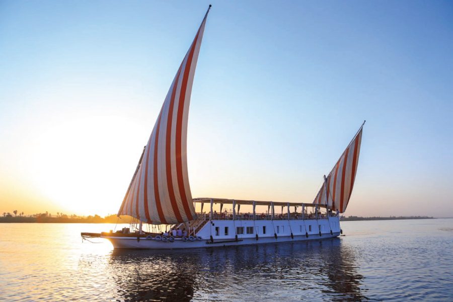 Une Croisière en Dahabyia , un grand voilier égyptien blanc avec deux voiles rayées de rouge et blanc, navigue sur le Nil au coucher du soleil. Le soleil, à gauche, se reflète sur l'eau et le ciel est un mélange de bleu clair et de teintes jaunes et roses. Les rives du fleuve sont visibles à l'arrière-plan.