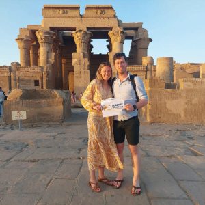 Un jeune couple, la femme vêtue d'une robe longue jaune et l'homme d'une chemise bleue, se tiennent debout devant le temple de Kom Ombo. Ils sourient et tiennent une pancarte blanche de l'agence de voyage "Alagabi Travel & Tours". Le temple en arrière-plan est illuminé par la lumière du soleil couchant, et d'autres personnes se promènent au loin.