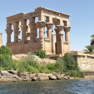Une vue latérale du Kiosque de Trajan sur l'île de Philae, montrant la structure en pierre antique avec ses colonnes et ses murs en ruine. L'île est couverte de végétation et un palmier se dresse près du kiosque. Le bâtiment est situé sur un rivage rocheux au bord de l'eau.